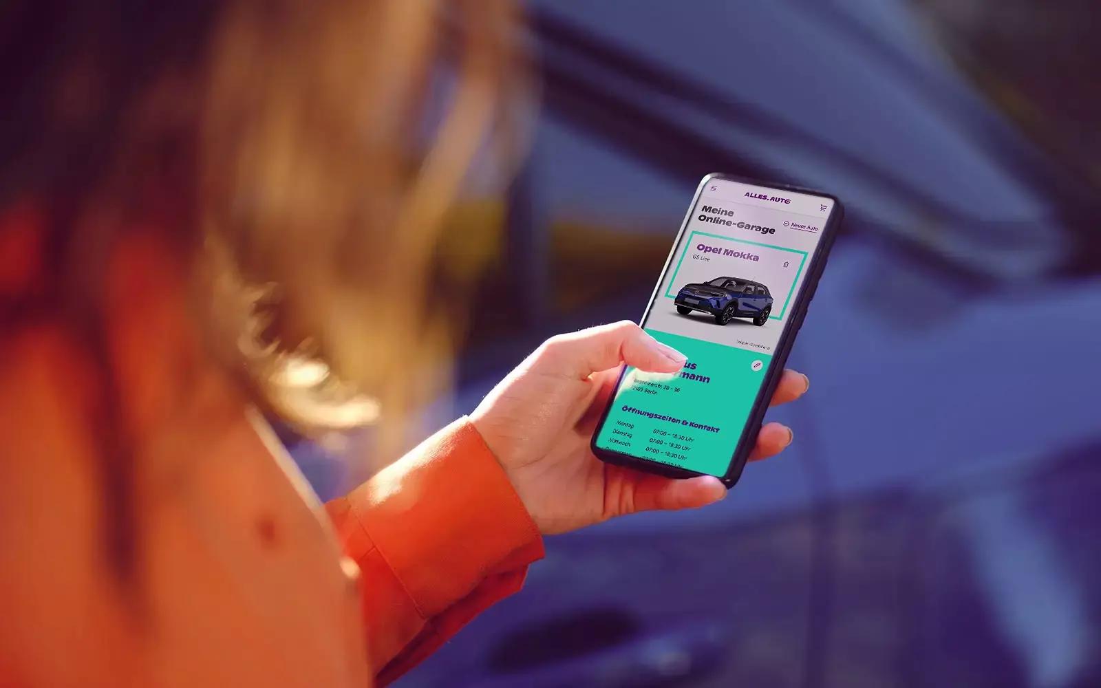 Frau will ihr Auto verkaufen und betrachtet auf einem Smartphone die Online-Garage mit einem blauen Opel Mokka.